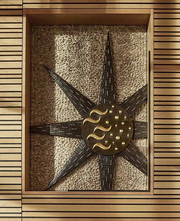 A central disc with wavy and circular gold-leafed elements on a dark glass circles with 7 rays emanting all on a buff-colored pebble mosaic field