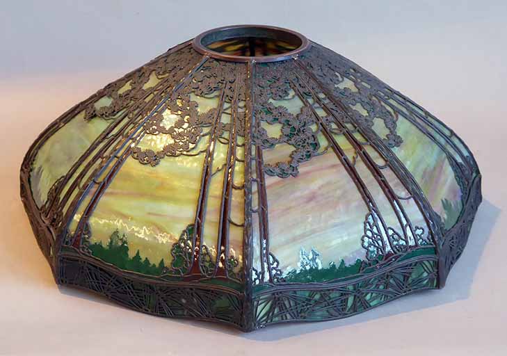 In natural (reflected) light: a curved glass lamp with a yellow/orange/blue main color with an metal overlay of evergreen trees and a green skirt overlaid with abstract curving lines