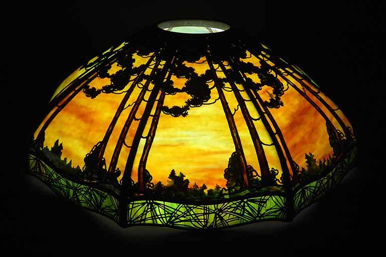 An illuminated curved glass lamp with a yellow/orange/blue main color with an metal overlay of evergreen trees and a green skirt overlaid with abstract curving lines