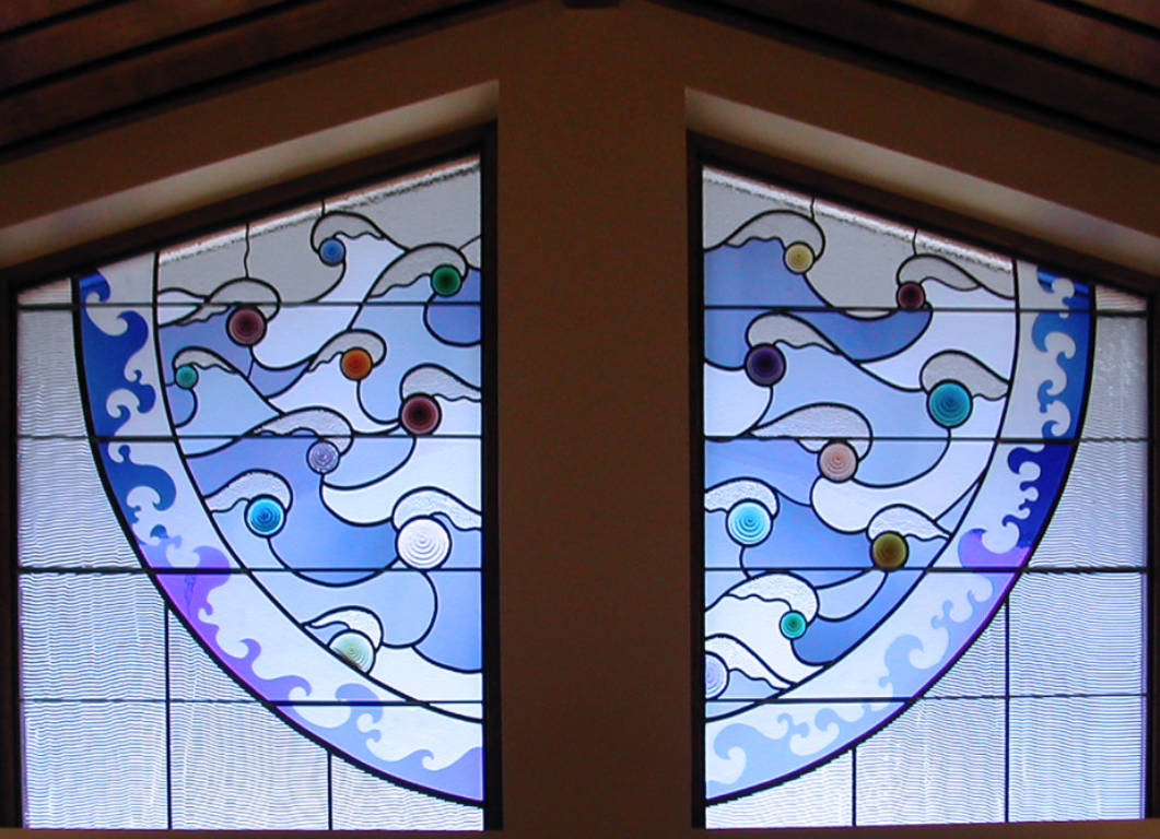 Two windows pointed at the center with the lower half of a circle filled with wave shapes encircling variously colored circles, all surrounded by a circle of etched wave shapes with clear ripple glass extending to the left, right and bottom edges of the opeinings