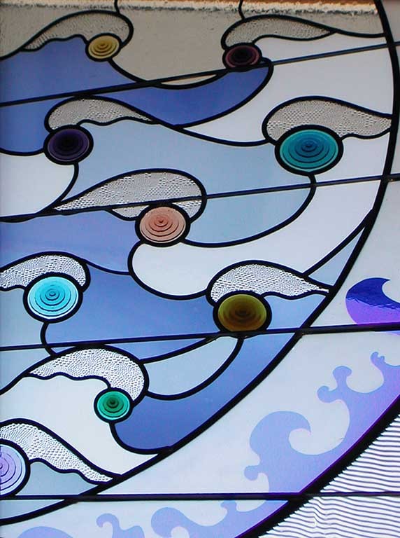 Detail shows the fused glass circles in an array of colors capping white, light blue and darker blue wave shapes bordered by an area of etched wave shapes